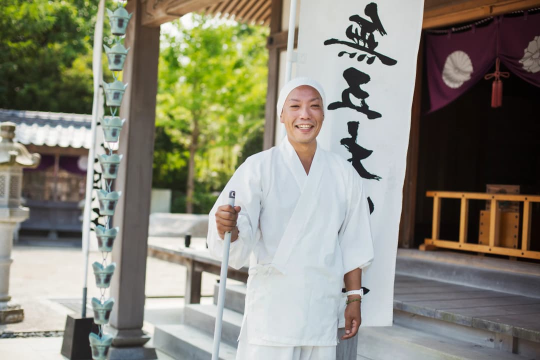 Buddhist monk wearing white robe and cap standing outside a temple, smiling at camera